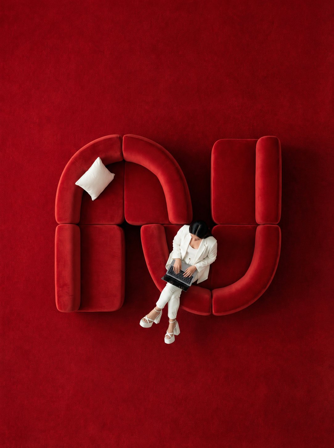 A woman in white working on a laptop on a red velvet sofa, seen from above